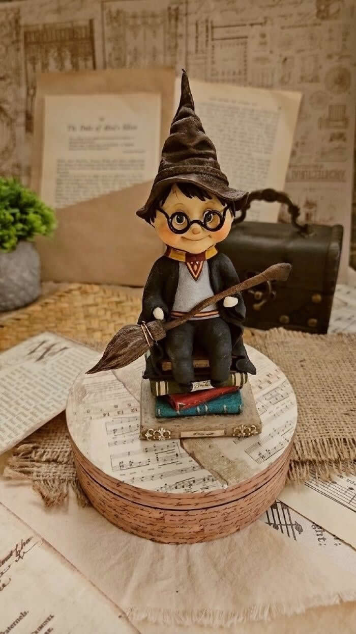 A handcrafted miniature wizard boy ornament sits on a stack of tiny books, holding a broomstick and wearing round glasses and a pointed hat, displayed on a vintage-style tabletop with sheet music, old papers, and a small suitcase in the background.
