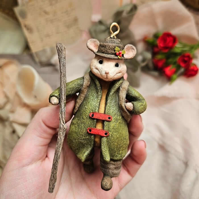 A handcrafted miniature mouse ornament made from cotton wool is dressed in a moss-green coat, tiny brown hat, and rustic boots, holding a walking stick while standing in a hand against a soft vintage-style background with candles, fabric, and red roses.