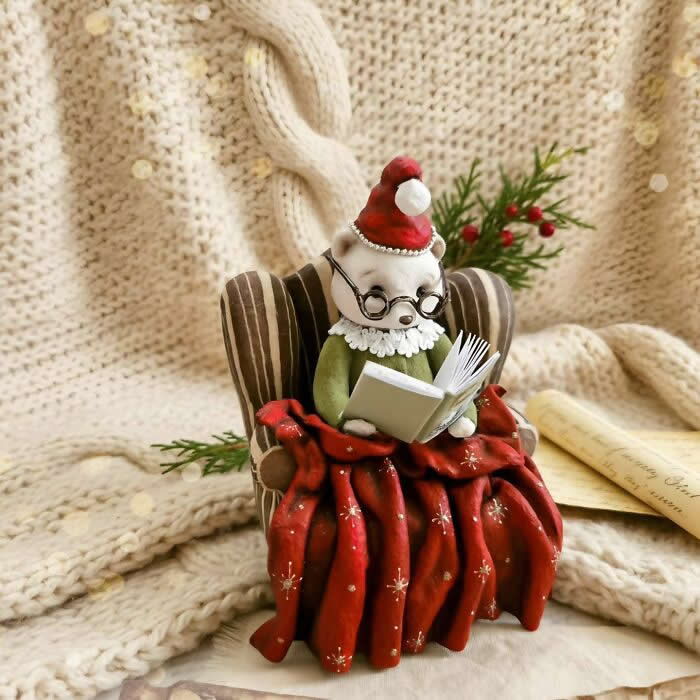 A handcrafted cotton wool mouse ornament sits in a striped armchair reading a tiny book, dressed in a green outfit, lace collar, round glasses, and a red Santa hat, wrapped in a red patterned blanket against a cozy knitted winter backdrop.