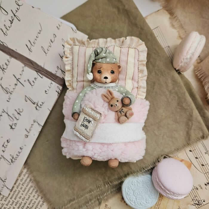 A handcrafted cotton wool bear ornament lies tucked into a tiny pink bed, wearing mint-green polka-dot pajamas and a matching nightcap, cuddling a small bunny toy and holding a miniature bedtime book, surrounded by vintage papers and pastel macarons.