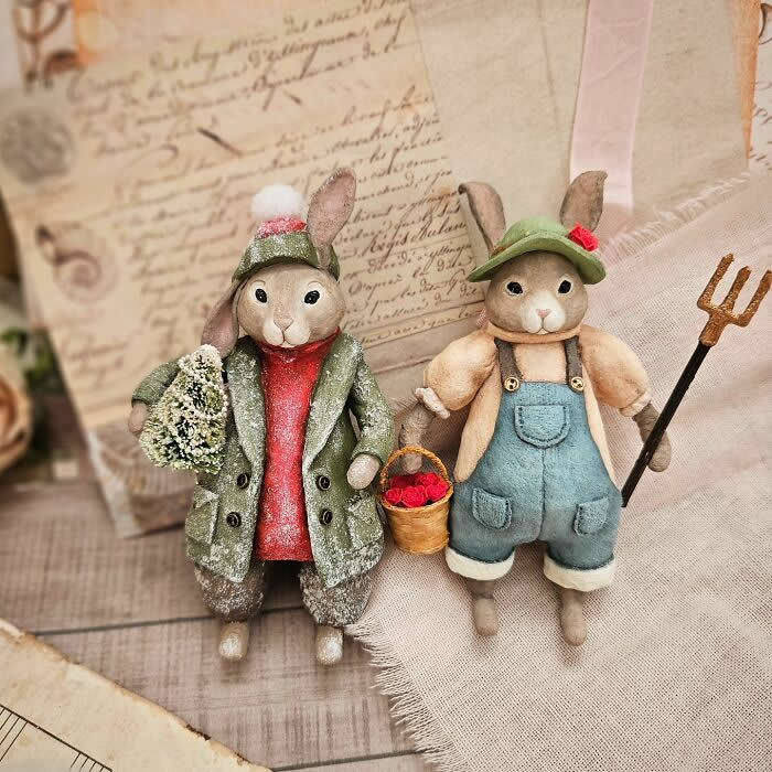 Two handcrafted cotton wool rabbit ornaments stand side by side in a vintage-style scene&mdash;one dressed in a frosted winter coat holding a tiny tree, the other in blue overalls with a basket of red berries and a small pitchfork&mdash;set against soft fabric and antique paper backgrounds.