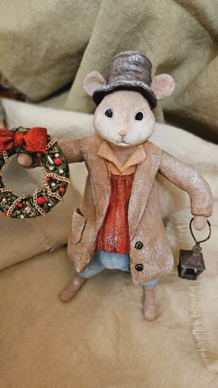 A handcrafted cotton wool mouse ornament stands on soft neutral fabric, dressed in a frosted beige coat and tall top hat, holding a festive wreath in one hand and a tiny lantern in the other, styled in a vintage holiday aesthetic.