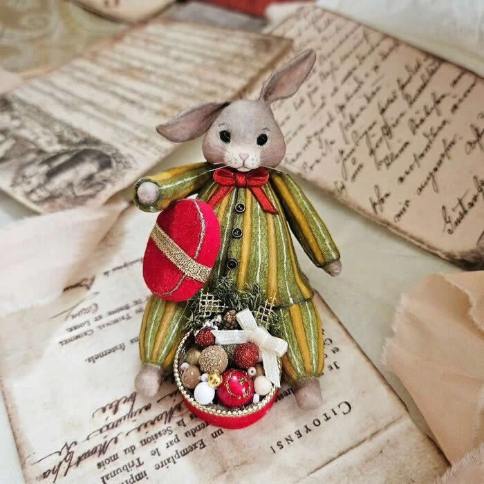 A handcrafted cotton wool rabbit ornament in a striped green-and-gold outfit stands on vintage paper, holding a large red ornament and a tiny basket filled with miniature festive decorations, finished with a bright red bow at the neck.