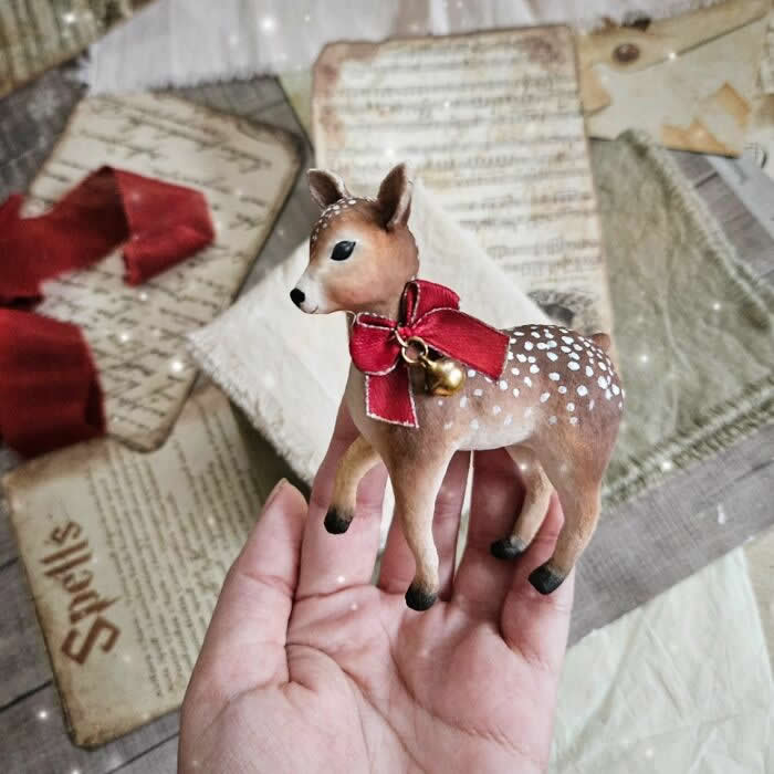 A handcrafted cotton wool deer ornament with delicate white spots stands in a hand against a vintage paper background, wearing a large red ribbon with a tiny gold bell, evoking a soft and festive woodland holiday charm.