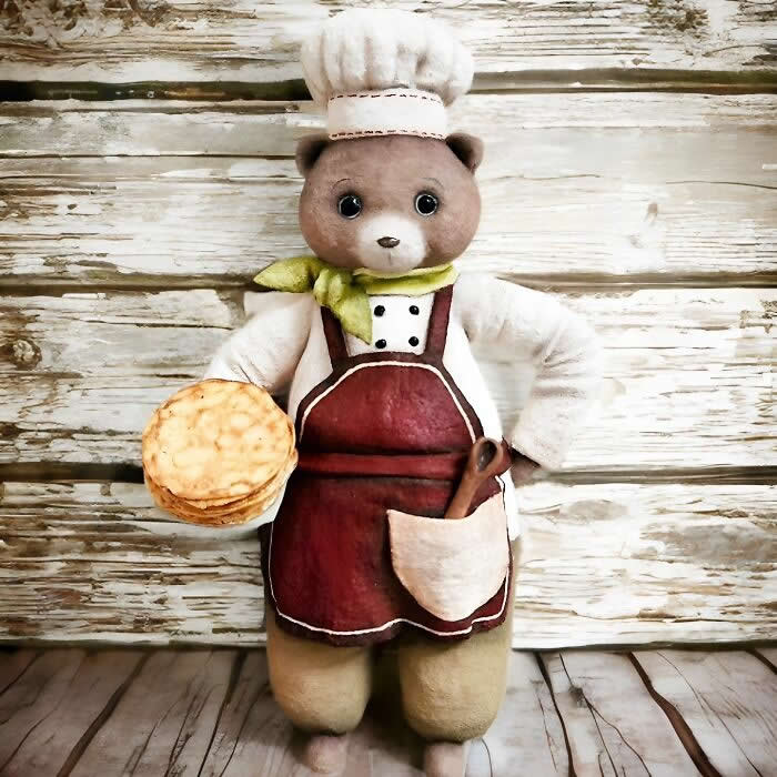 A handcrafted cotton wool bear ornament dressed as a baker stands against a rustic wooden backdrop, wearing a white chef&rsquo;s hat, green scarf, and burgundy apron while holding a stack of golden pastries, with a wooden spoon tucked into the apron pocket.