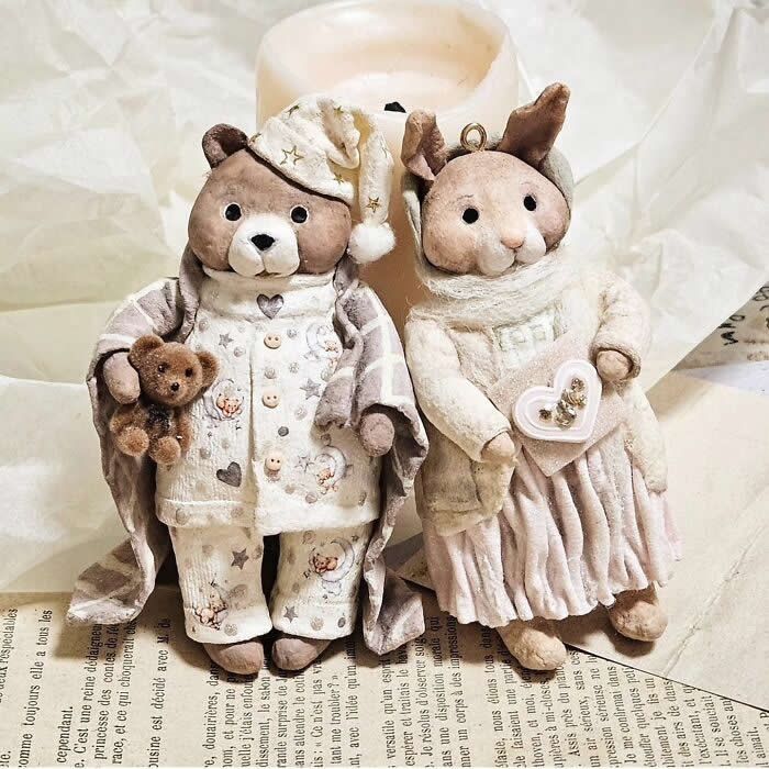 Two handcrafted cotton wool ornaments stand side by side on vintage paper in a cozy holiday setting: a teddy bear in cream pajamas and a nightcap holding a tiny teddy, and a rabbit in a soft white sweater and pleated skirt holding a heart-shaped keepsake.