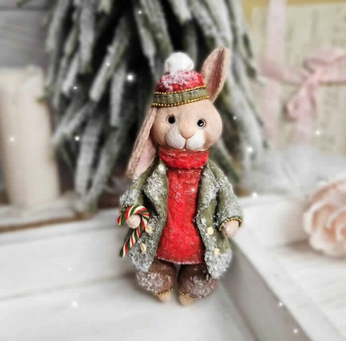 A handcrafted cotton wool rabbit ornament stands in a snowy holiday scene, dressed in a frosted green coat, bright red scarf, and knit winter cap, holding a tiny candy cane beside a blurred Christmas tree and soft candlelit background.
