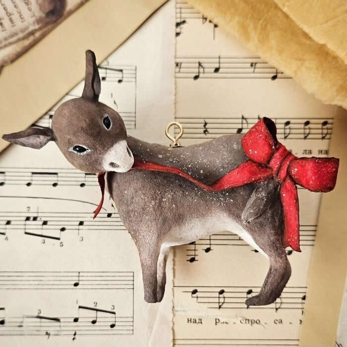 A handcrafted cotton wool reindeer ornament is posed against vintage sheet music, turning its head gently to the side while wearing a long red scarf tied in a dramatic bow, with softly dusted details that give it a wintry, storybook charm.