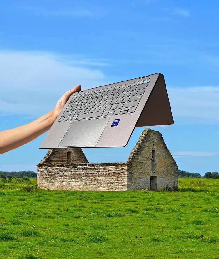 A hand holds an open laptop in the foreground, perfectly aligned with an old stone building in a grassy field so the laptop appears to become the roof of the house through creative forced perspective.