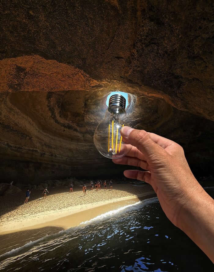 A hand holds a clear light bulb in front of a cave opening, aligning it so the bright sunlight pouring into the sea cave looks like the bulb is glowing above a sandy beach and small people below.