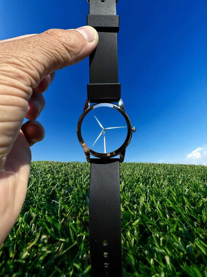 A hand holds a black wristwatch against a bright blue sky and grassy field, aligning the watch face with a distant wind turbine so the turbine blades appear to become the hands of the watch in a playful forced perspective illusion.
