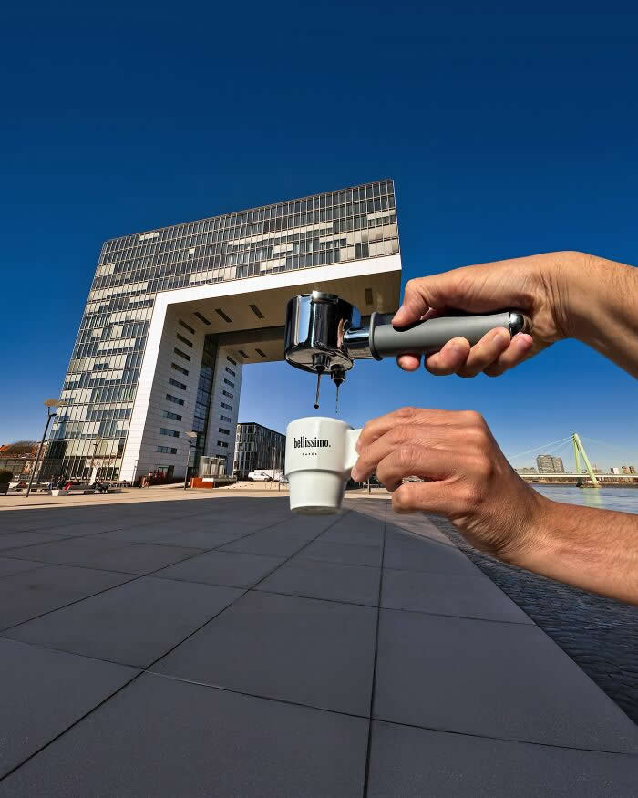 A hand holds an espresso portafilter above a small coffee cup, aligned with a large modern arch-shaped building in the background so the building appears to pour coffee into the cup through a clever forced perspective illusion.