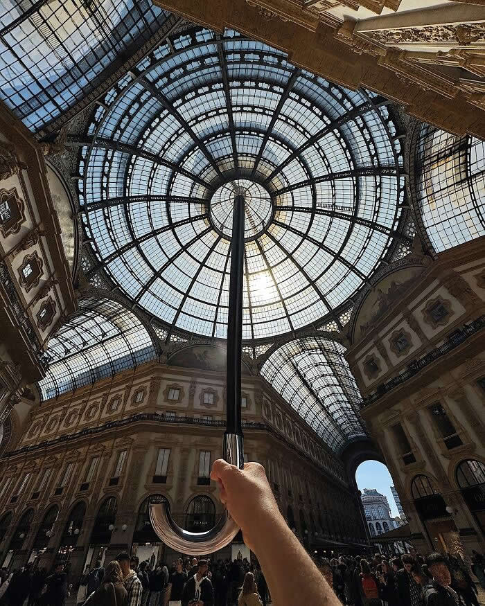 Inside a grand glass-roofed arcade, a hand holds a closed umbrella upward, perfectly aligned with the circular dome so it looks like the umbrella is supporting the entire ceiling in a dramatic forced perspective composition.