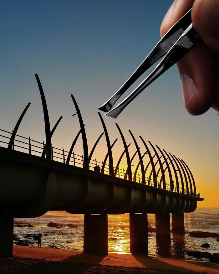 A hand holds a pair of tweezers above a seaside pier at sunset, aligned so the tweezers appear to pluck one of the curved metal beams from the structure in a clever forced perspective illusion.