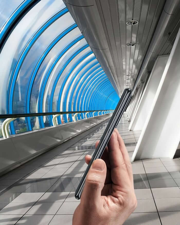 A hand holds a slim smartphone inside a curved glass walkway, aligned so the phone appears to fold shut around the tunnel, turning the arched passage into part of a playful forced perspective illusion.