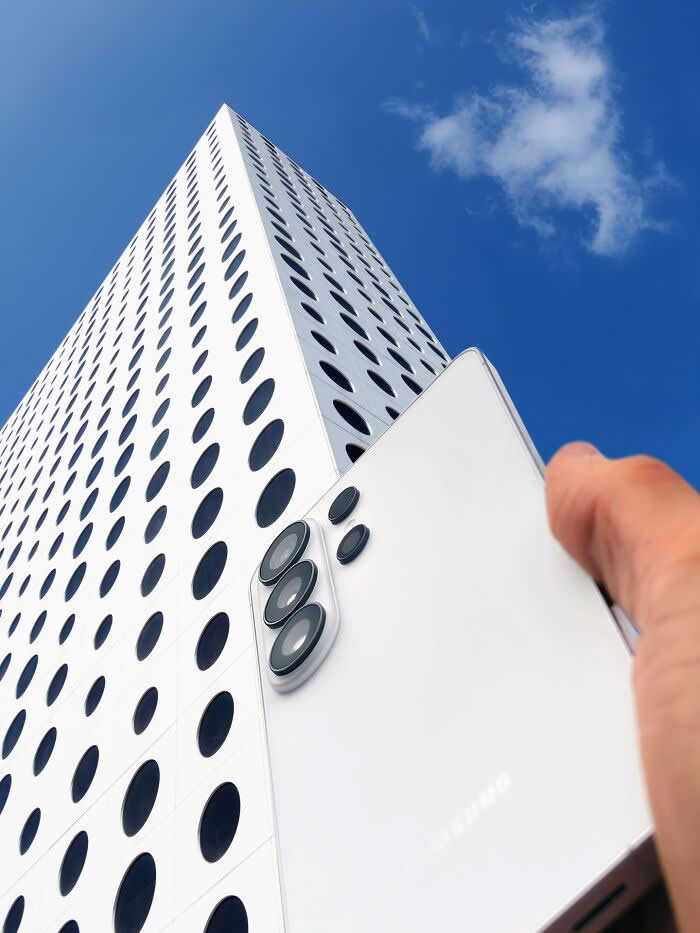 A hand holds a white smartphone in front of a tall white building with circular black windows, aligning the phone&rsquo;s rear camera lenses with the building&rsquo;s dotted fa&ccedil;ade to create a playful forced perspective illusion.