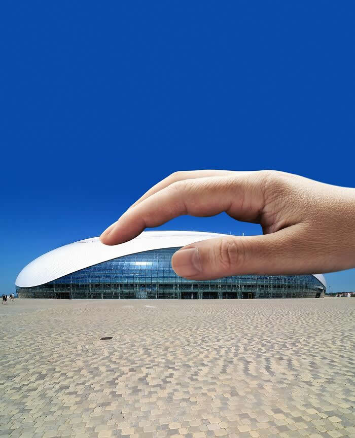 A hand hovers above a sleek oval-shaped modern building, aligned so the fingers appear to gently pinch or press the curved white roof against a deep blue sky in a clever forced perspective composition.