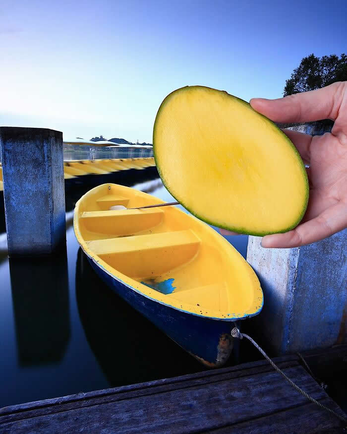 A hand holds half a mango in the foreground, perfectly aligned with a yellow boat floating in calm water so the fruit appears to become the boat&rsquo;s upper half in a playful forced perspective illusion.