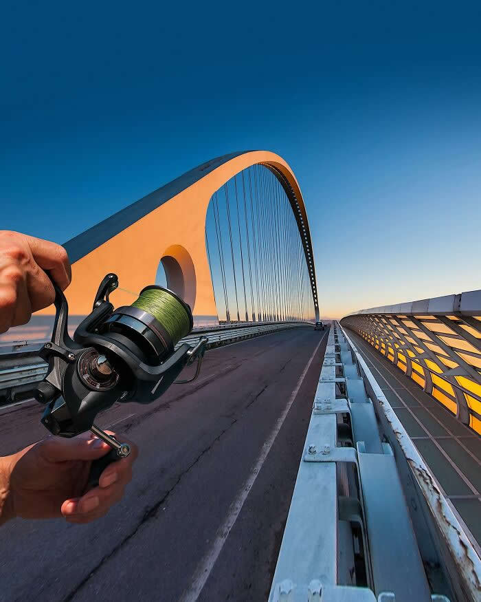 A hand holds a fishing reel in the foreground, aligned with a large arched bridge at sunset so the bridge appears to be threaded through the reel like a fishing line in a playful forced perspective illusion.