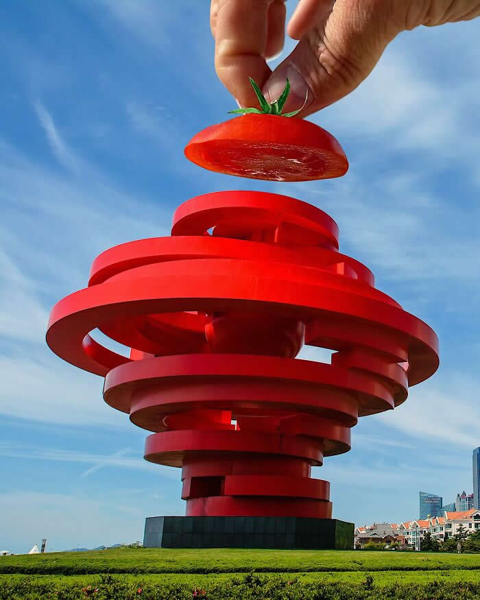 A hand holds a sliced tomato above a large red spiral sculpture, aligned so the tomato appears to become the top layer of the structure in a bold forced perspective illusion against a blue sky.