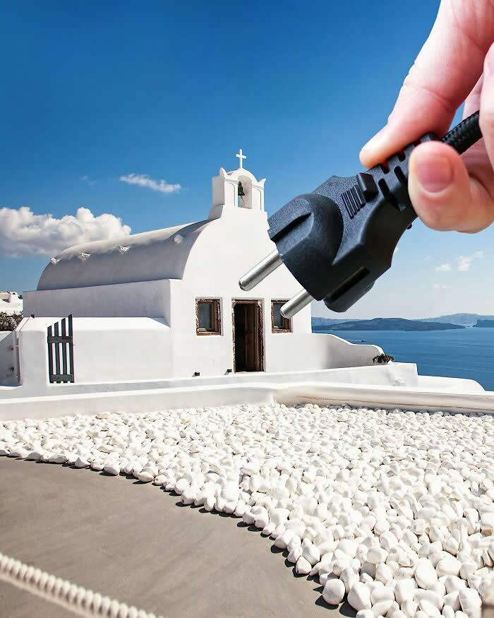 A hand holds a black electrical plug in the foreground, aligned with a white seaside chapel so the plug appears to connect directly into the building, creating a playful forced perspective illusion against the blue sky and sea.