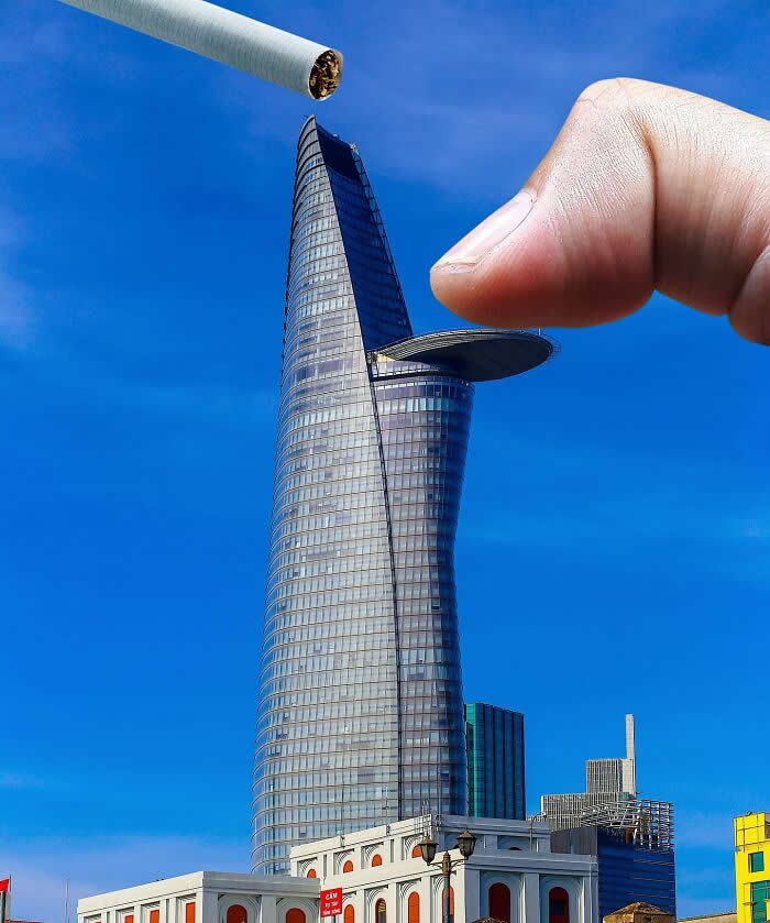 A hand pinches the circular helipad jutting from a tall glass skyscraper while a cigarette hovers above the tower, making it look as if the building is being used like an ashtray in a clever forced perspective illusion.