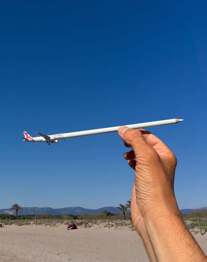 A hand holds a white pencil in the foreground, perfectly aligned with a plane flying across a clear blue sky so the aircraft appears to be attached to the pencil like it&rsquo;s being drawn in a playful forced perspective illusion.
