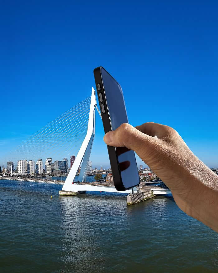 A hand holds a black smartphone in the foreground, aligned with a white cable-stayed bridge so the bridge&rsquo;s tall central pylon appears to emerge from the phone like a sleek extension in a playful forced perspective illusion.