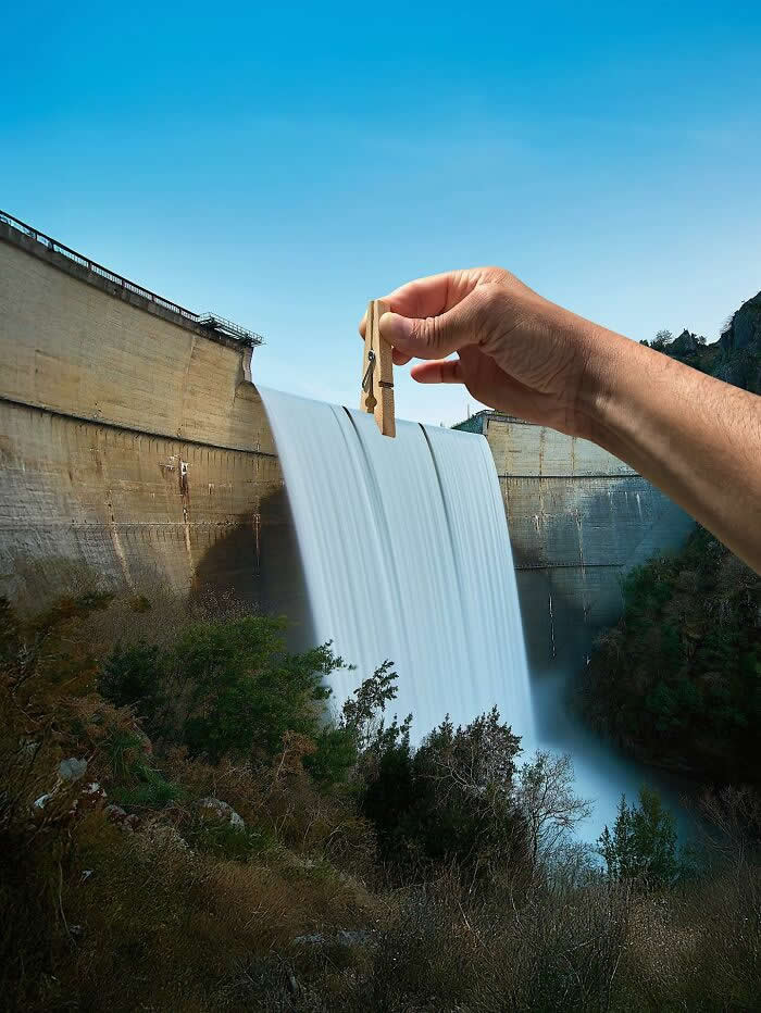A hand holds a wooden clothespin above a dam spillway, aligned so the pin appears to clip the rushing sheet of water as it pours over the edge in a playful forced perspective illusion.