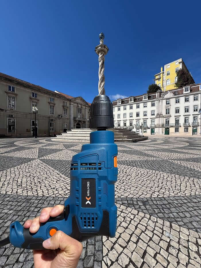 A hand holds a blue power drill in the foreground, aligned with a tall stone column in a city square so the monument appears to become the drill bit in a playful forced perspective illusion.