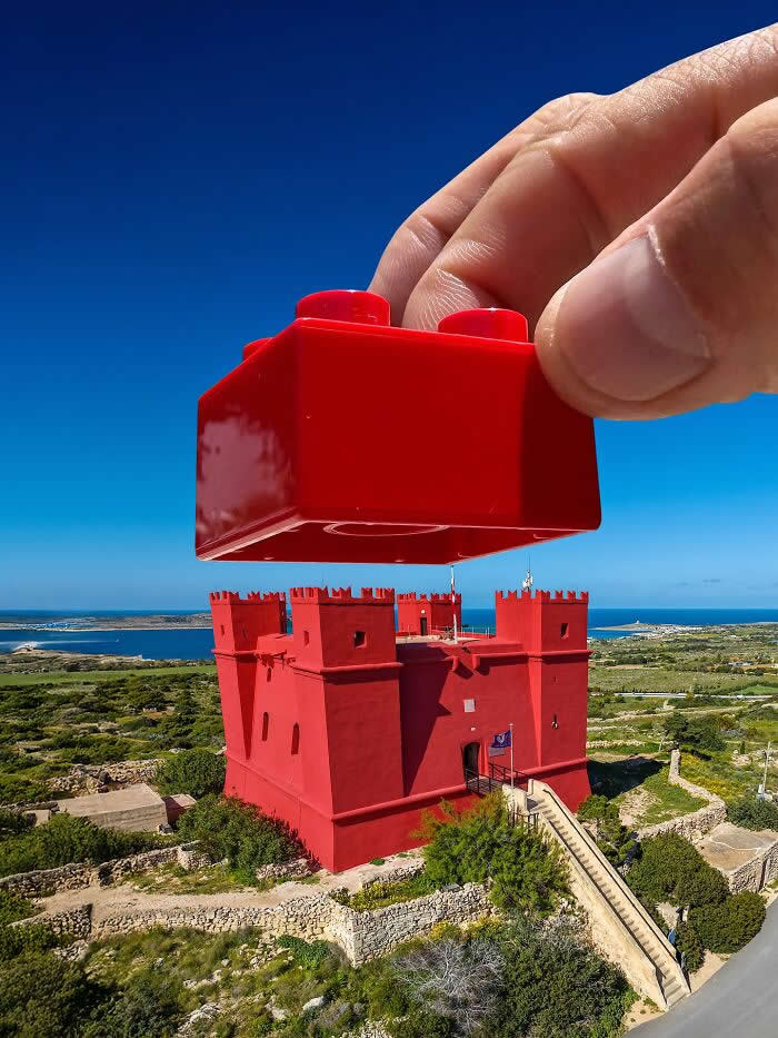 A hand holds a red toy building block above a bright red fortress, aligned so the block appears to become the castle&rsquo;s missing top piece in a playful forced perspective illusion against a vivid blue sky.