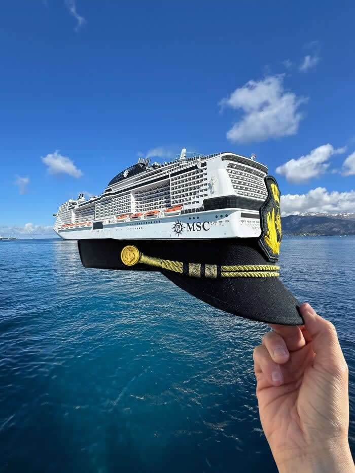 A hand holds a black captain&rsquo;s hat in the foreground, perfectly aligned with a large white cruise ship on calm blue water so the ship appears to be wearing the hat in a playful forced perspective illusion.