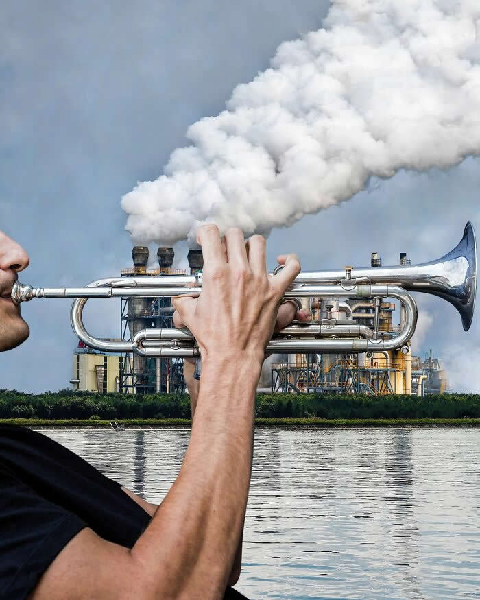 A man holds a silver trumpet in the foreground, aligned with an industrial plant in the background so the factory&rsquo;s thick white smoke looks like it&rsquo;s blasting out of the trumpet in a playful forced perspective illusion.
