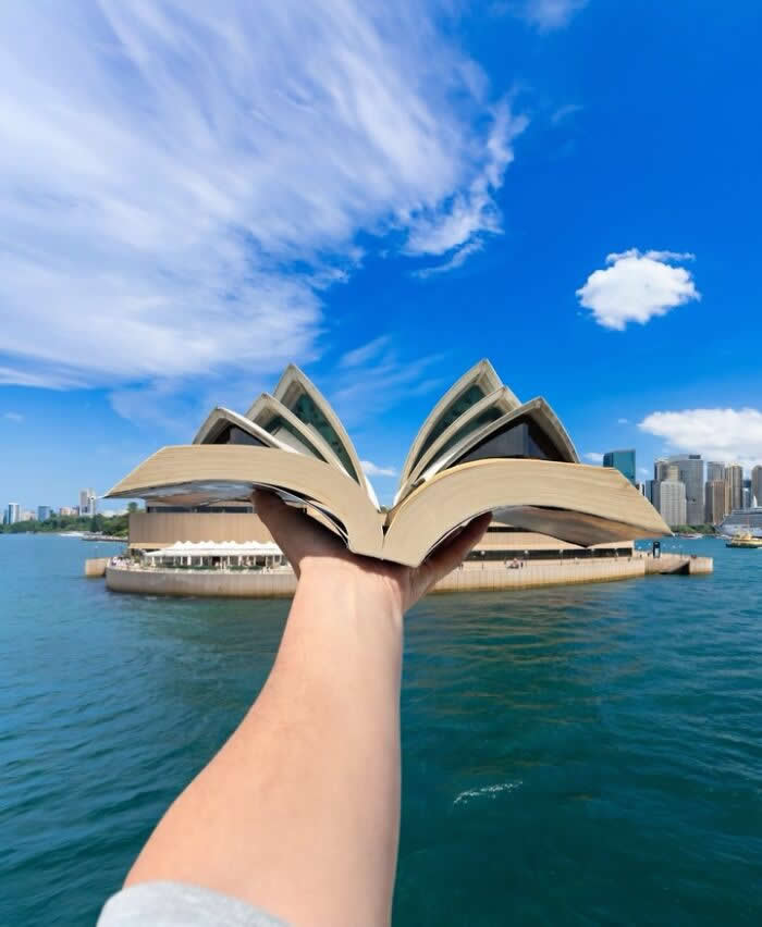 A hand holds an open book in the foreground, perfectly aligned with the Sydney Opera House so the building&rsquo;s iconic sail-like roofs appear to rise from the book&rsquo;s pages in a playful forced perspective illusion.