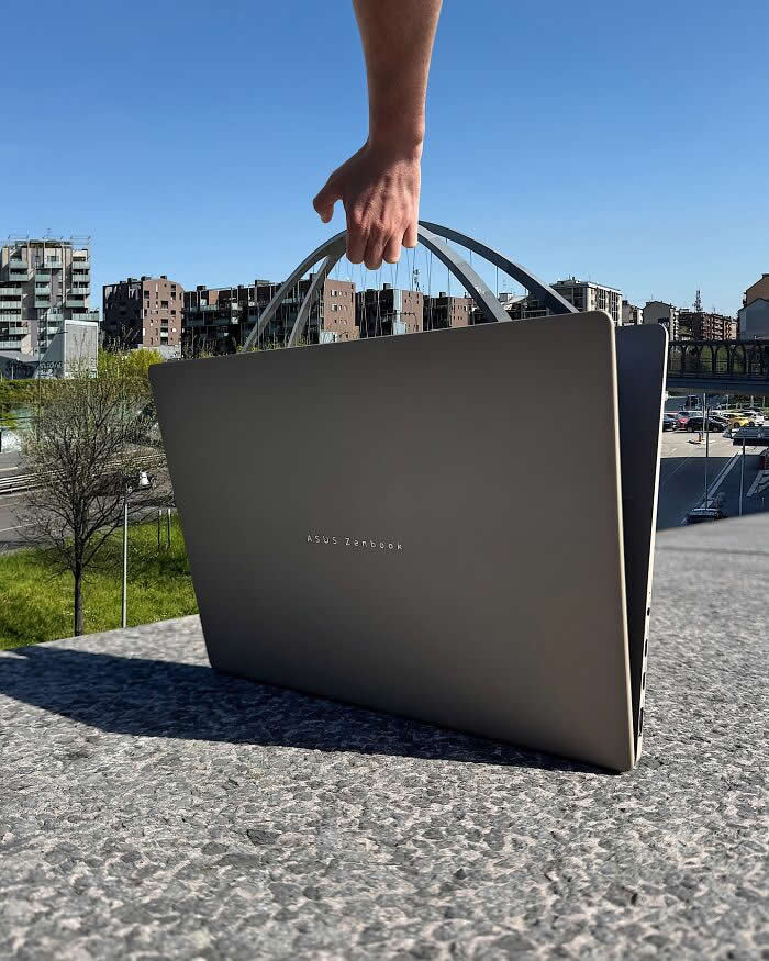 A hand grips the arched handle of a bridge in the background, aligning it with a closed laptop in the foreground so the bridge appears to become the laptop&rsquo;s carrying handle in a playful forced perspective illusion.