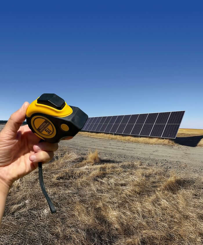 A hand holds a yellow tape measure in the foreground, aligned with a long row of solar panels in a dry open field so the panels appear to extend directly from the measuring tape in a playful forced perspective illusion.