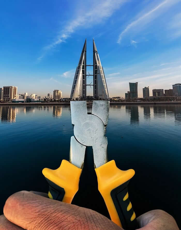 A hand holds a pair of yellow-handled pliers in the foreground, aligned with the twin sail-shaped Bahrain World Trade Center towers so the pliers appear to grip the skyscrapers in a playful forced perspective illusion above calm waterfront reflections.