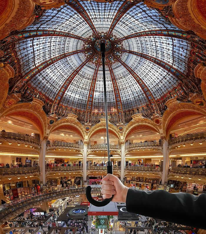 Inside an ornate shopping gallery with a stained-glass dome, a hand holds an umbrella upward, perfectly aligned so the umbrella appears to support the entire ceiling in a playful forced perspective illusion.