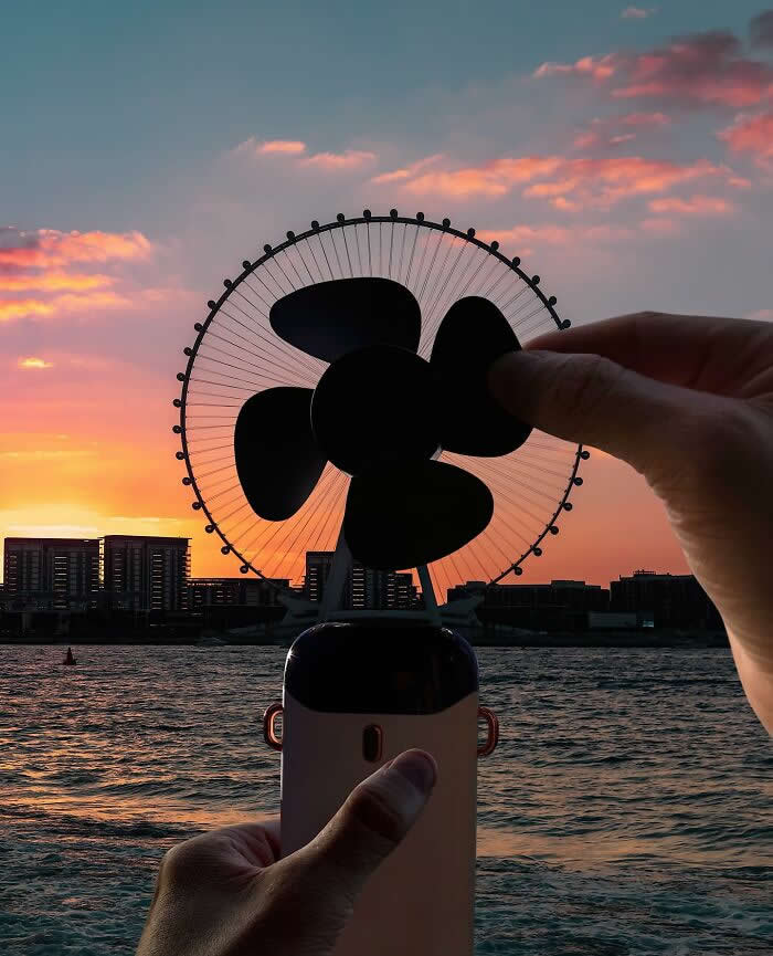 At sunset by the water, a hand holds a small portable fan aligned with a giant Ferris wheel in the background, making the wheel look like the fan&rsquo;s oversized spinning blades in a clever forced perspective composition.