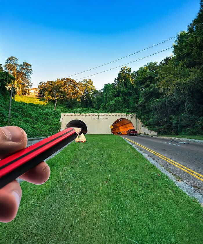 A hand holds a red pencil in the foreground, aligned with a pair of road tunnels so the pencil appears to slide directly into one tunnel like a sharpened tip in a playful forced perspective illusion.