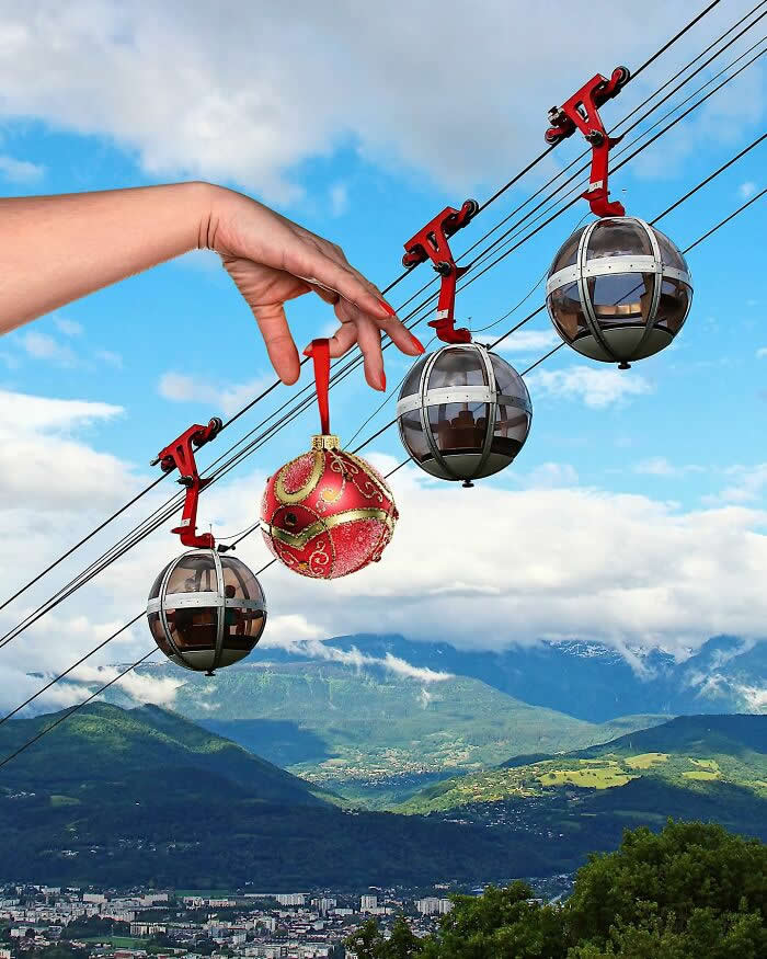 A hand holds a red Christmas ornament in front of a cable car line, aligning it between the gondolas so it looks like the ornament is hanging from the same cable among the cabins in a clever forced perspective scene above a mountain landscape.