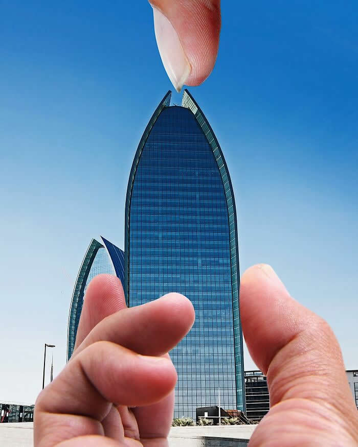 Two hands frame a tall curved glass skyscraper against a clear blue sky, aligned so the fingertips appear to pinch the pointed top of the building in a playful forced perspective illusion.