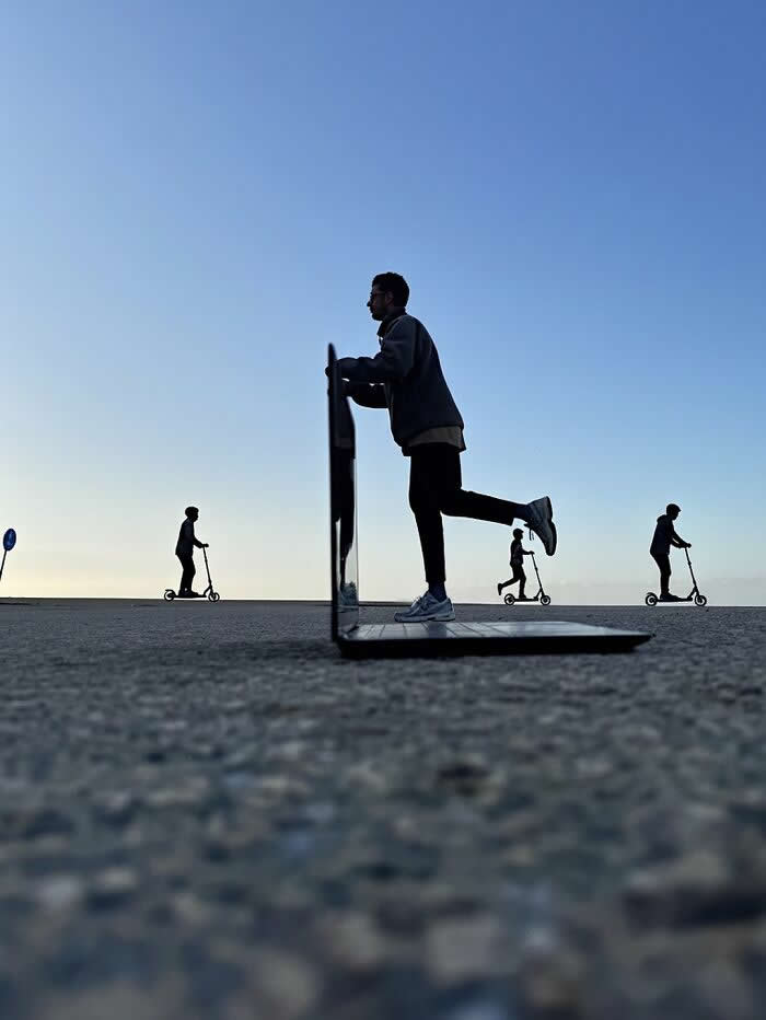 A person appears to ride a scooter through the screen of an open laptop placed on the ground, while other scooter riders move across the horizon in the background, creating a playful forced perspective illusion.