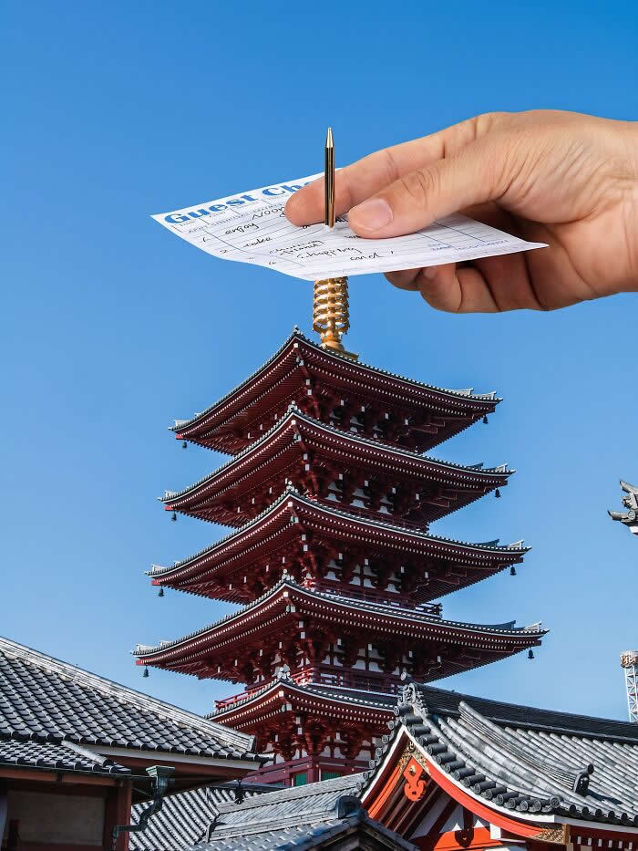 A hand holds a guest check and pen above a traditional five-story pagoda, aligned so the pen appears to be inserted into the pagoda&rsquo;s golden spire, creating a playful forced perspective illusion against a clear blue sky.