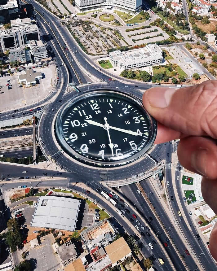 A hand holds a black wristwatch above an aerial view of a large traffic roundabout, aligning the watch face with the circular road so the roundabout appears to become part of the watch in a playful forced perspective illusion.