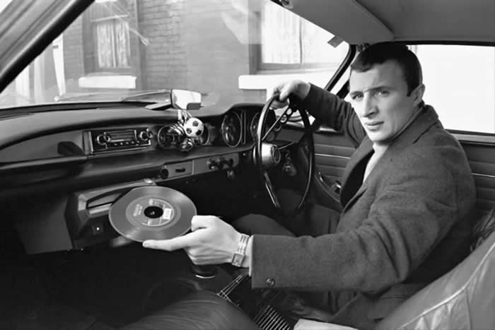 Manchester City player Mike Summerbee demonstrating the built- in record player of his new Volvo P1800 (1967).