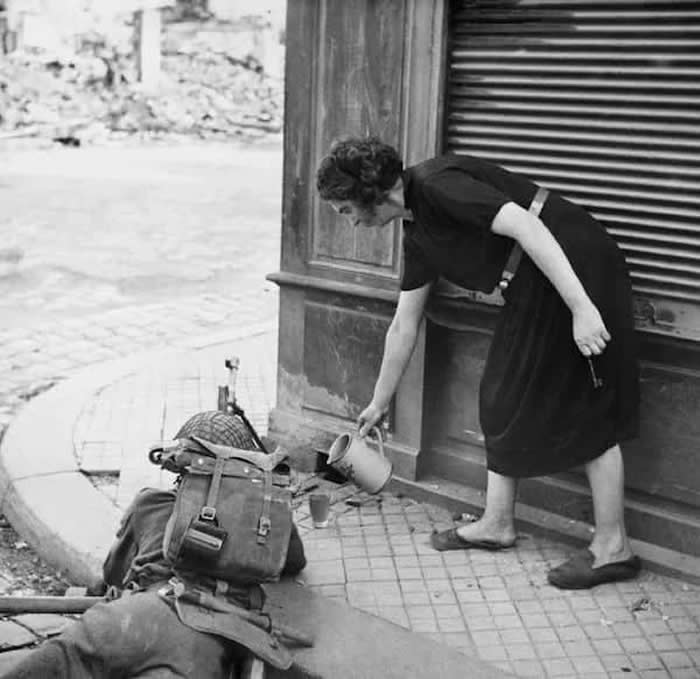 A French civilian woman pours a drink of cider for a British soldier with Bren gun in Lisieux, 1944