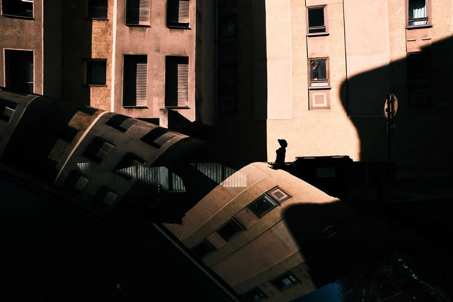 The Art of Perfect Timing: 34 Clever Street Photos by Emiliano Cuadrado That Turn Ordinary Scenes Into Visual Surprises 40 Silhouette of a lone person standing beside urban buildings, framed by bold sunlight, deep shadows, and mirrored reflections that create a layered, abstract street photography composition.
