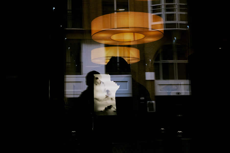 The Art of Perfect Timing: 34 Clever Street Photos by Emiliano Cuadrado That Turn Ordinary Scenes Into Visual Surprises 41 Silhouette of a person seen through a reflective glass window, layered with warm hanging lights and architectural reflections, creating an abstract and cinematic street photography scene.