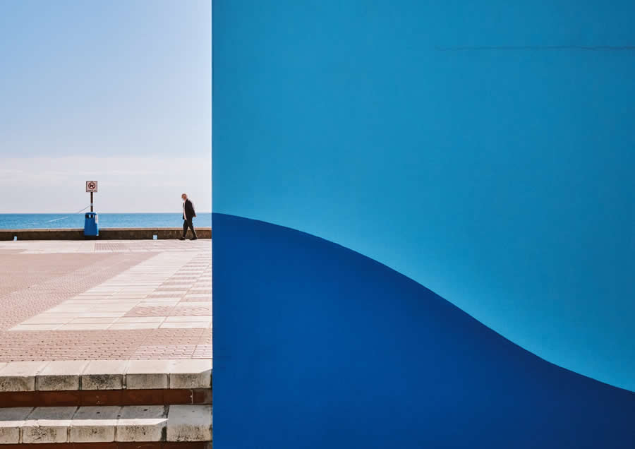 The Art of Perfect Timing: 34 Clever Street Photos by Emiliano Cuadrado That Turn Ordinary Scenes Into Visual Surprises 48 A lone pedestrian walks beside the sea, partially framed by a large blue wall that splits the composition, creating a minimalist street photography scene with bold colors and negative space.